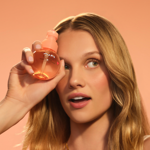 Woman holding Sunny Buns Hair & Body Mist beside her face against a peach background