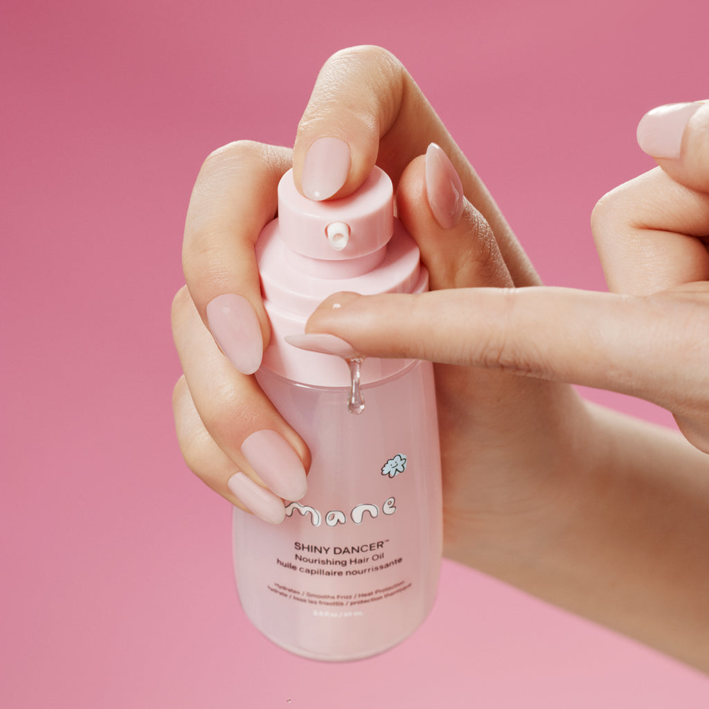 Hand holding a pink bottle of 'Shiny Dancer' nourishing hair oil against a pink background