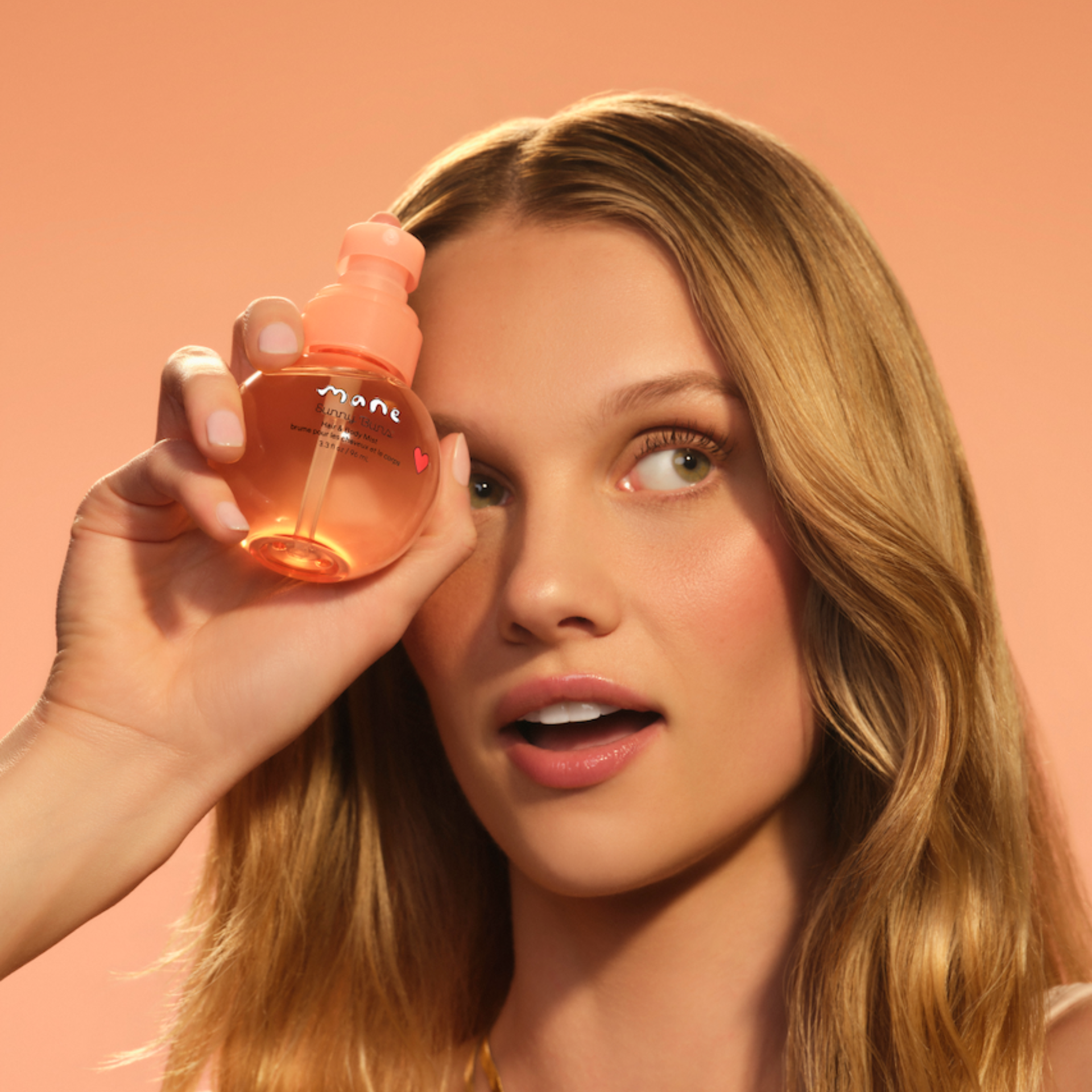 Woman holding Sunny Buns Hair & Body Mist beside her face against a peach background