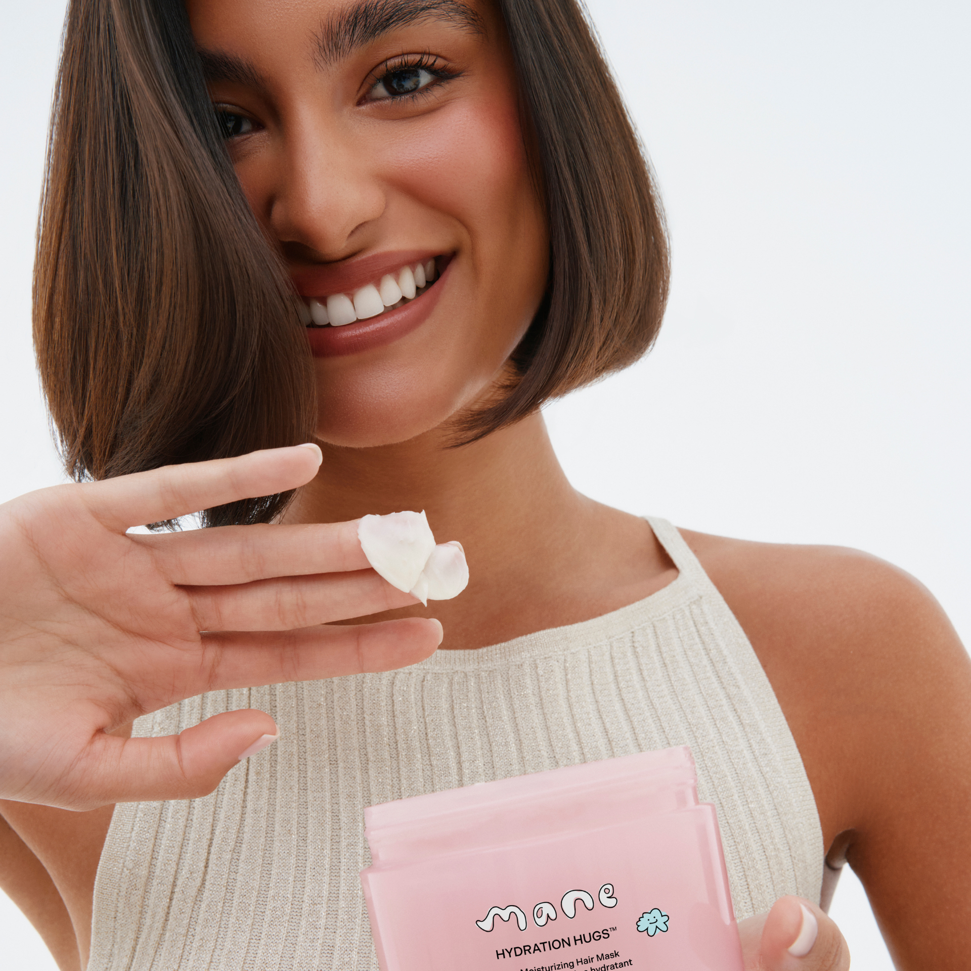 Woman holding a pink hair mask package with a small amount of product on her finger against a neutral background